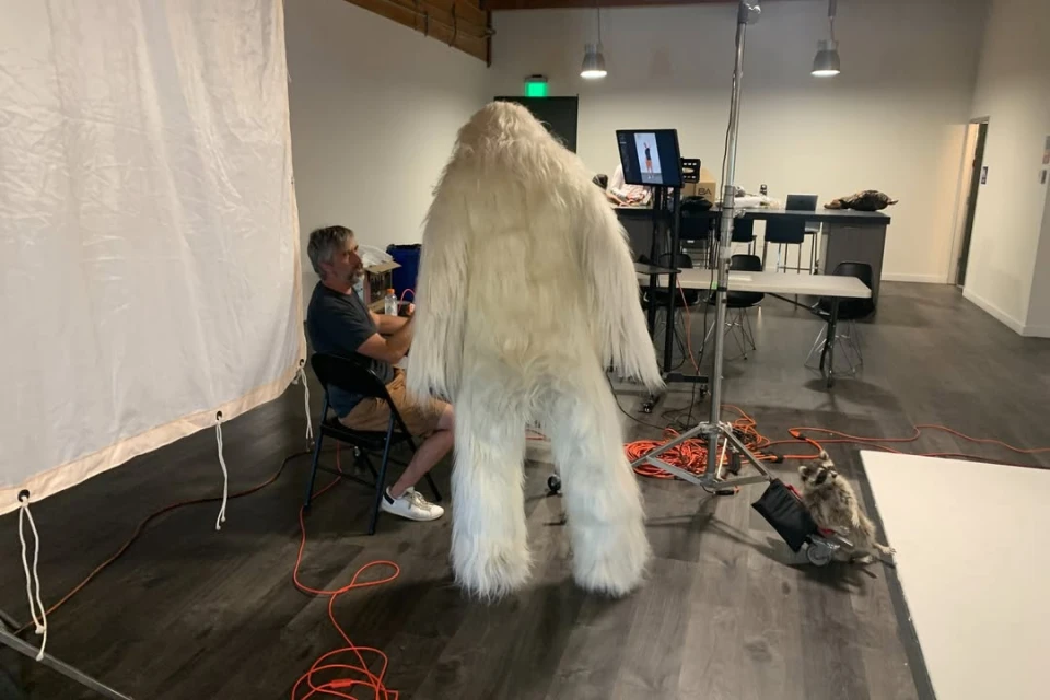 Man in a yeti costume stands in a studio with photographer, equipment, and white backdrop, ready for a creative shoot.